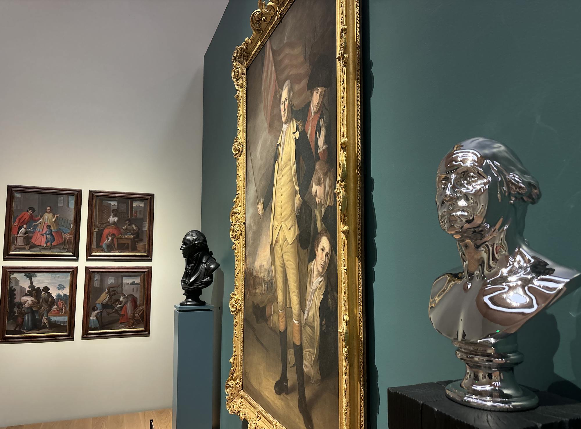A work of art depicting George Washington in a beige outfit hangs on a blue wall. The art is sandwiched by two busts, one black and one silver. The picture is taken at an angle, and a white wall is visible. On the wall hangs four colorful works of art.