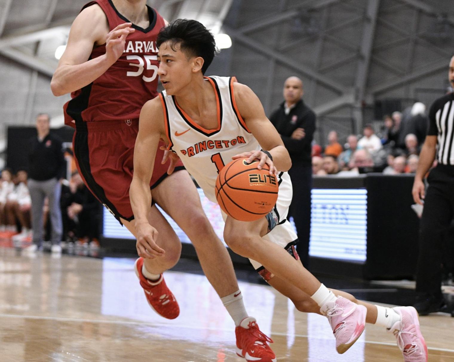 Basketball player in white Princeton jersey dribbles a basketball while defended by a basketball player in a red Harvard Jersey.