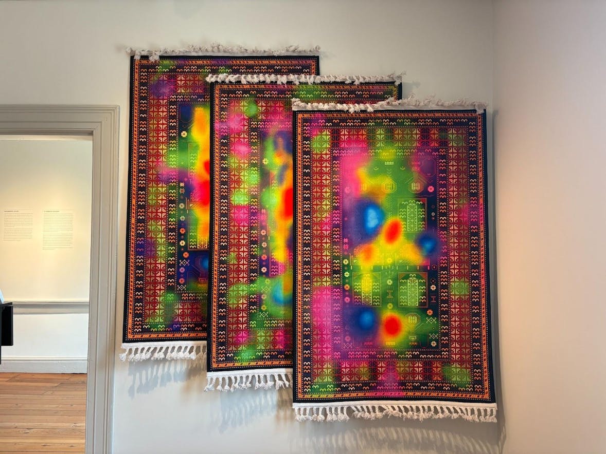Three carpets are hung against a white wall, overlapping and layering. The carpets have colorful neon hues of pink, green, yellow, red, and blue fading into one another. 