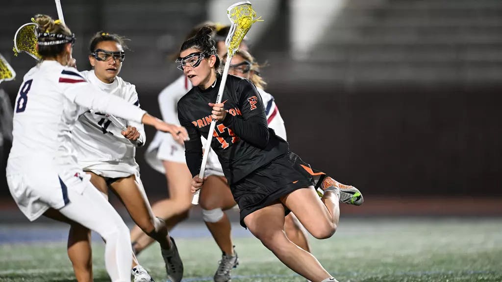 Woman in black lacrosse jersey carries the ball while guarded by three players in white jerseys.