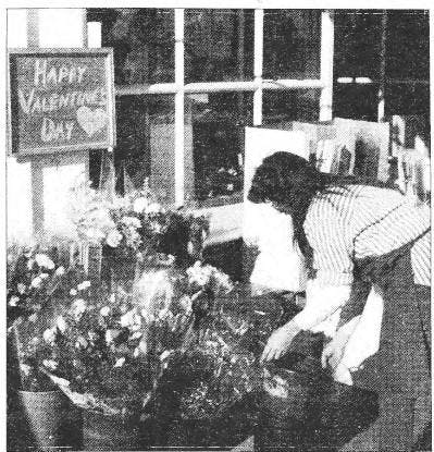 A black and white photograph of a person bending over and arranging bouquets of flowers outside of a storefront. A sign above the bouquets reads “Happy Valentine’s Day.”
