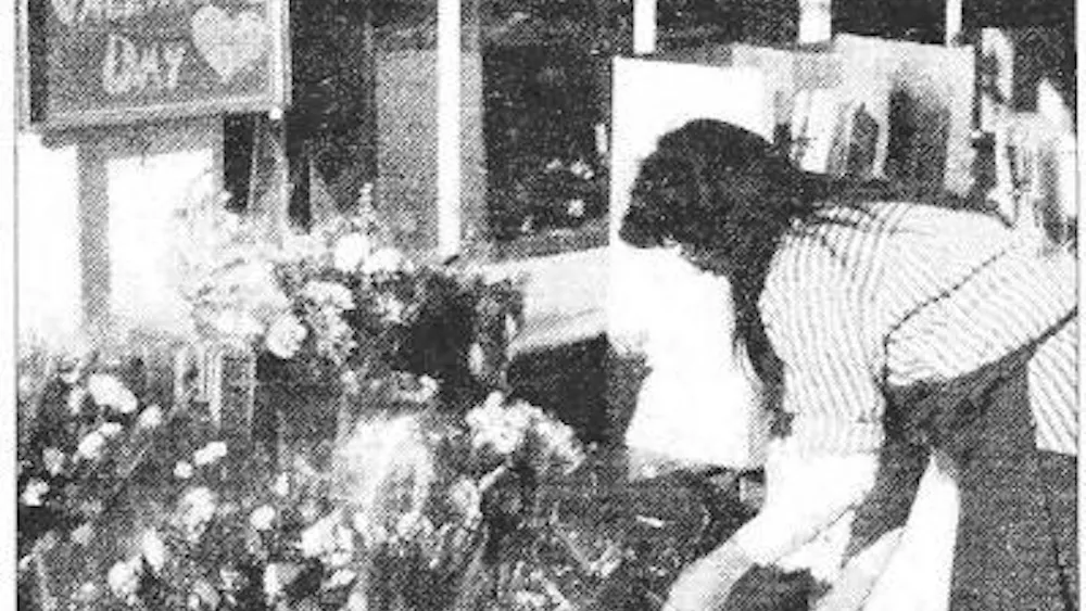 A black and white photograph of a person bending over and arranging bouquets of flowers outside of a storefront. A sign above the bouquets reads “Happy Valentine’s Day.”