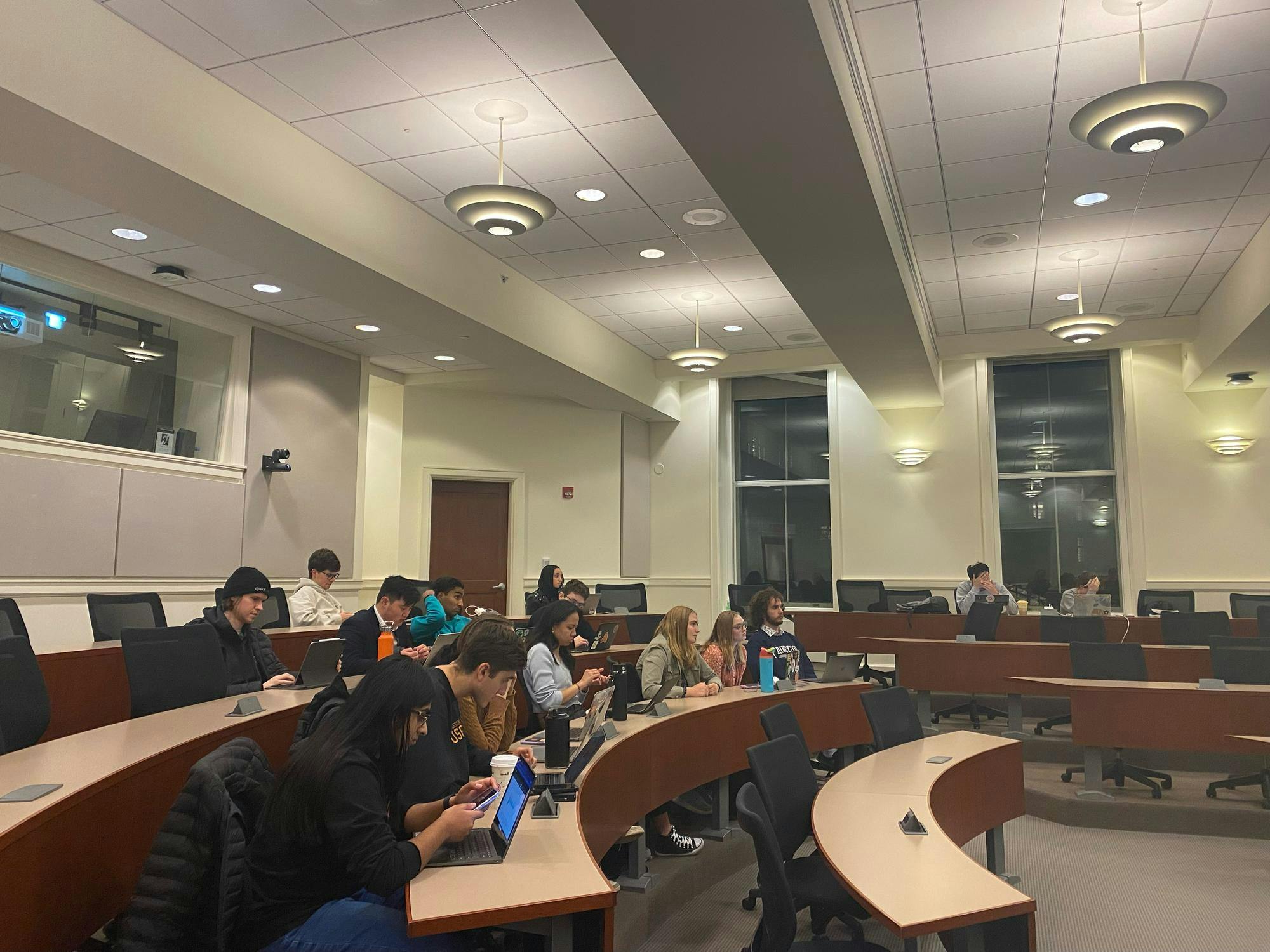 Students sit and type on their computers and phones in black spinning desk chairs in front of three large wooden tables.