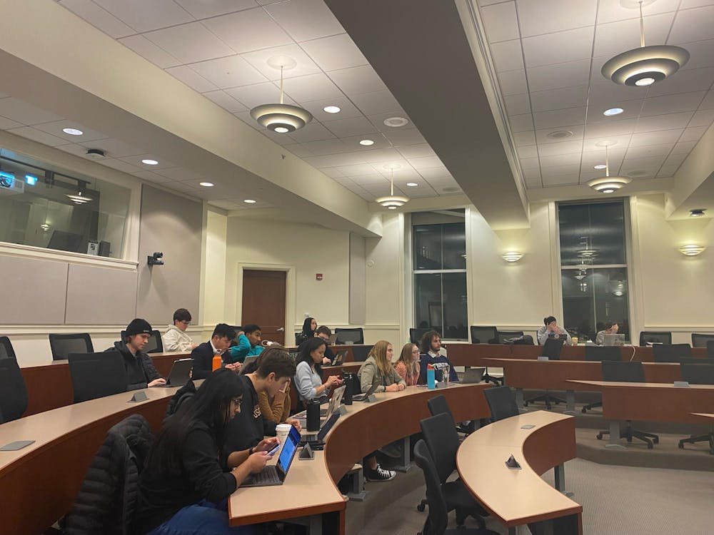 Students sit and type on their computers and phones in black spinning desk chairs in front of three large wooden tables.