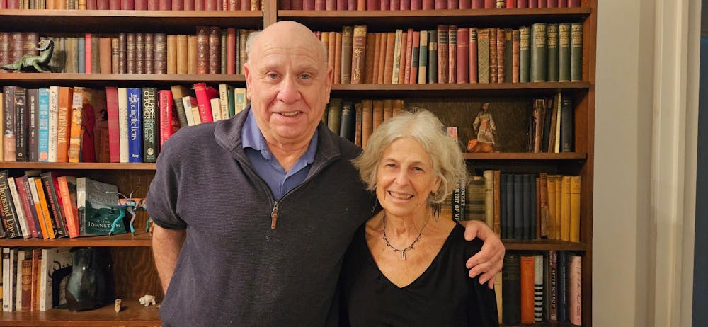 A man and a woman stand with their arms around each other in front of a bookcase full of books and trinkets. 