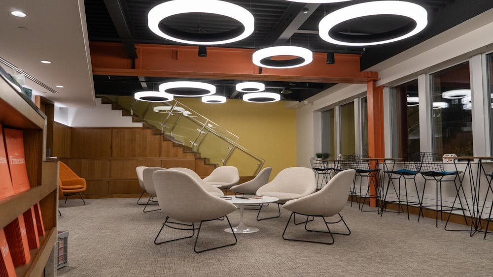 A modern lounge featuring circular ceiling lights, beige armchairs around a white coffee table, and a staircase with glass railings.