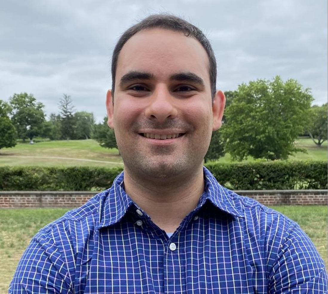 A man wearing a blue shirt stands smiling in front of a brick wall with a grassy green golf course in the background. 