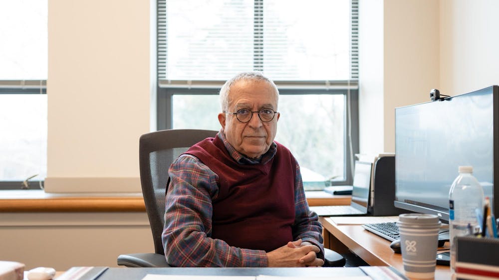 A older man with glasses and a red weather sitting facing the camera next to a computer