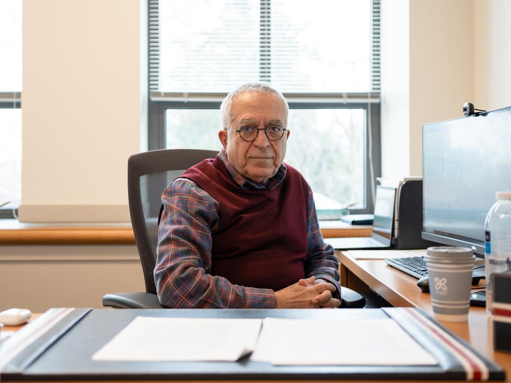 A older man with glasses and a red weather sitting facing the camera next to a computer