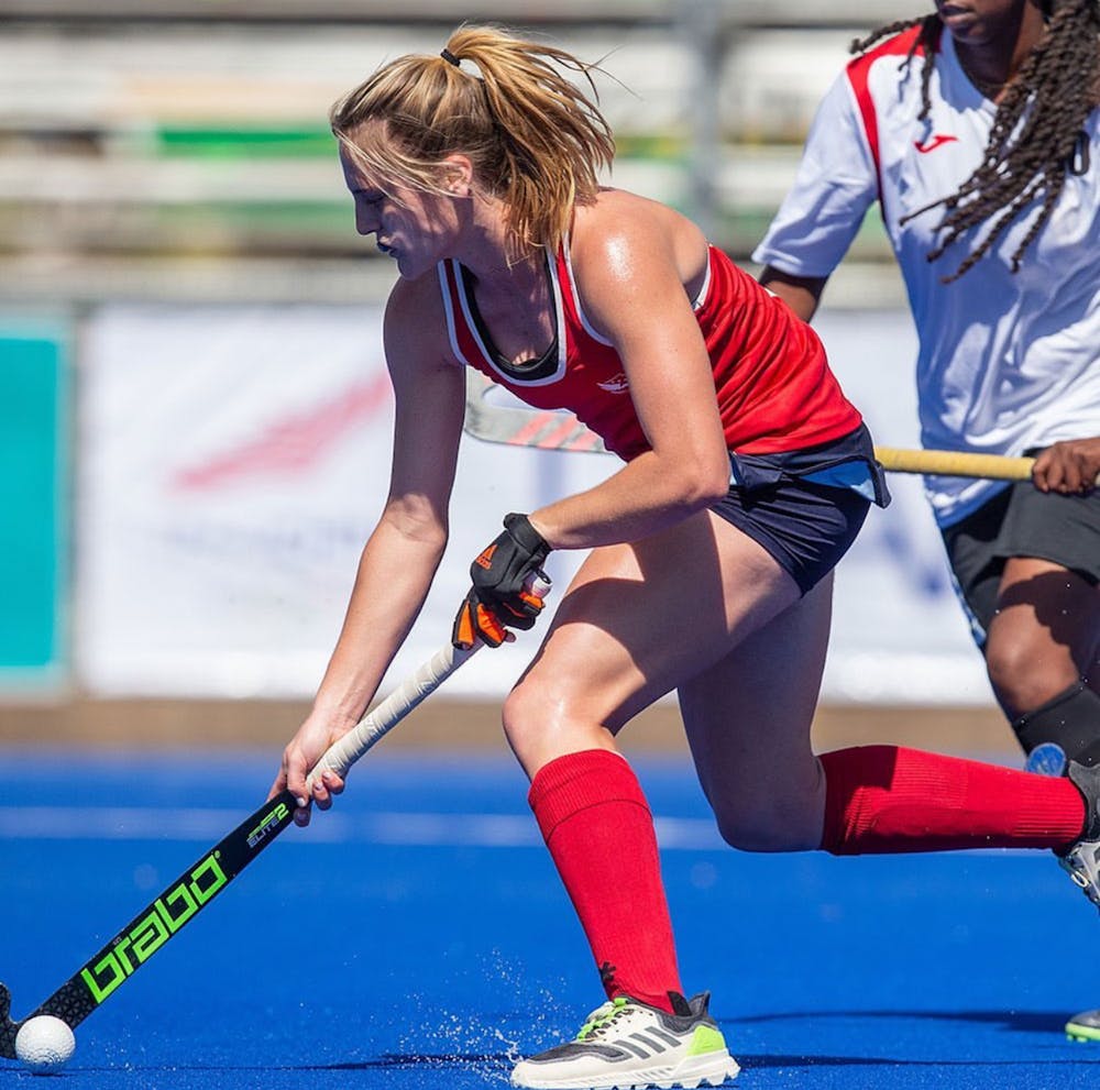 A woman holding a field hockey stick with another woman chasing her on a green pitch. 