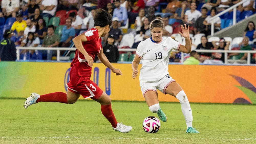 Two soccer players, one in a white uniform and another in a red uniform, run toward the ball on a green field.