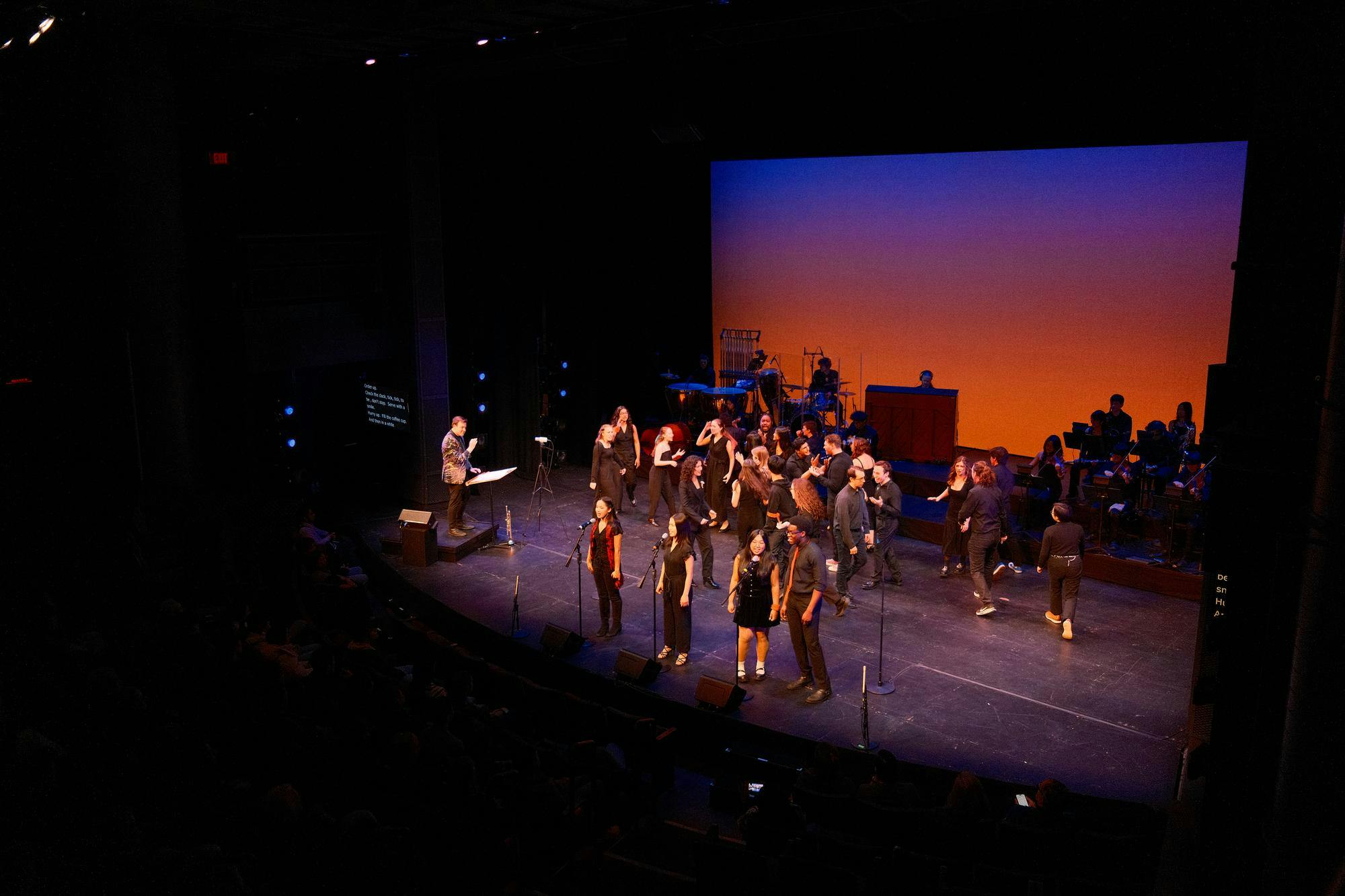 Student performers sing, play instruments, and move across a stage. 