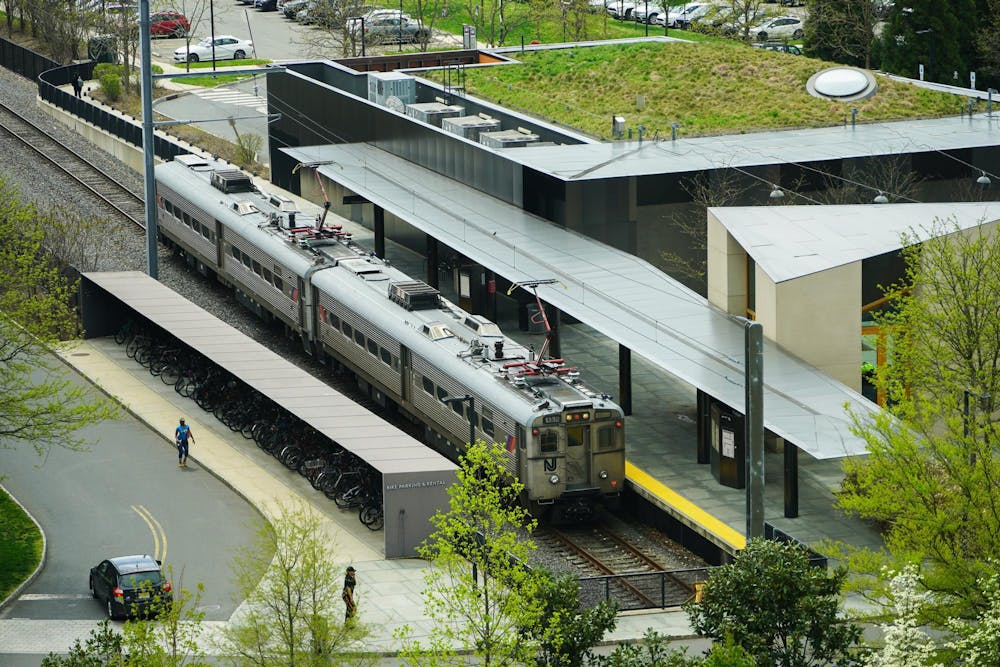 A silver train sits at a platform covered by a gray canopy. On the left, green trees.