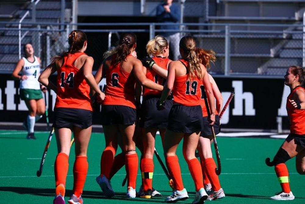 A group of field hockey players in orange and black uniforms celebrate on the field after a goal.