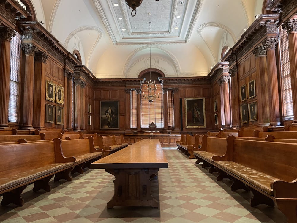 The picture shows the Princeton University Faculty Room in Nassau Hall. A long wooden table is at the center of the room. Brown wooden pews surround the table. Portraits line the walls, and a chandelier hangs from the ornate ceiling.