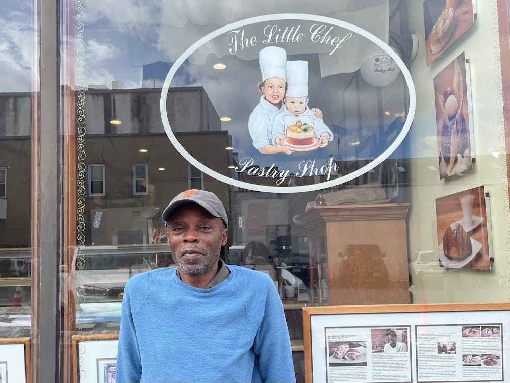 A man in a blue sweater and faded Princeton baseball cap stands in front of a pastry shop window, which features a logo with two children in chef outfits and white text that reads "The Little Chef Pastry Shop."