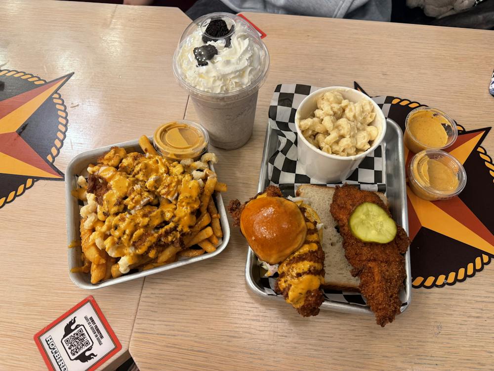 Multiple full sized dishes sit on a tan table. There are orange and light tan loaded fries. There is a combo meal with orange chicken, a slider with a light brown bun with orange colored chicken, and a side of yellow colored mac and cheese in a small white cup. There is also a light brown colored milkshake in a clear cup. There are also two yellow colored sauces in small transparent cups.