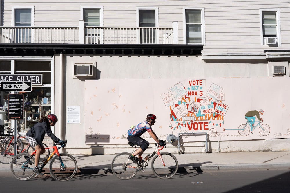 Two bikers on the left in foreground, with large tan wall behind that depicts signs that say "vote."