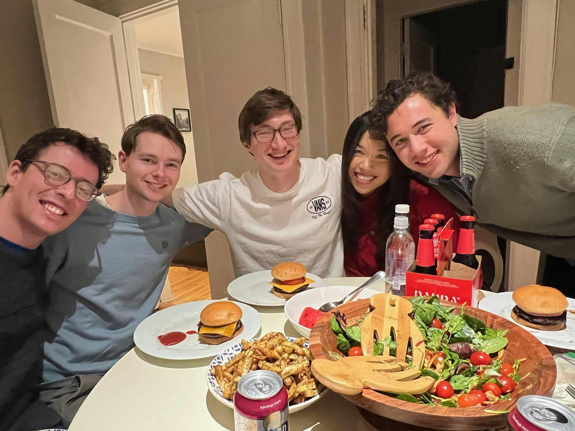 Five people smile with their arms around each other, seated at a circular table. Burgers, a large salad bowl, and a six-pack of drinks sit in front of them