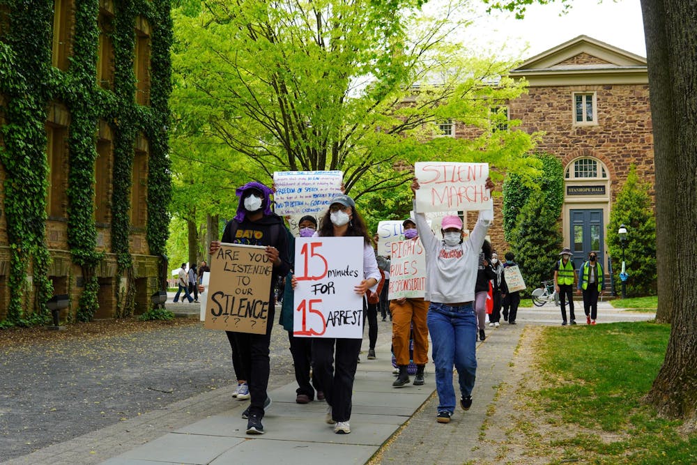 A group of students wearing hats and surgical masks marches past a large, ivy-colored building. They are holding signs that read, “15 minutes for 15 arrest,” “Are you listening to our silence,” “Silent March!” and “Rights Rules Responsibilities.”