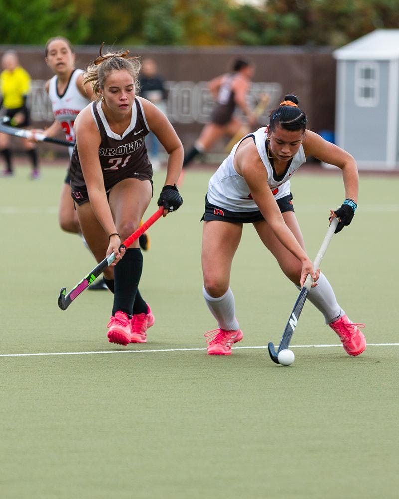 A woman in a white and orange jersey holding a field hockey stick, outcompeting her opponent. 