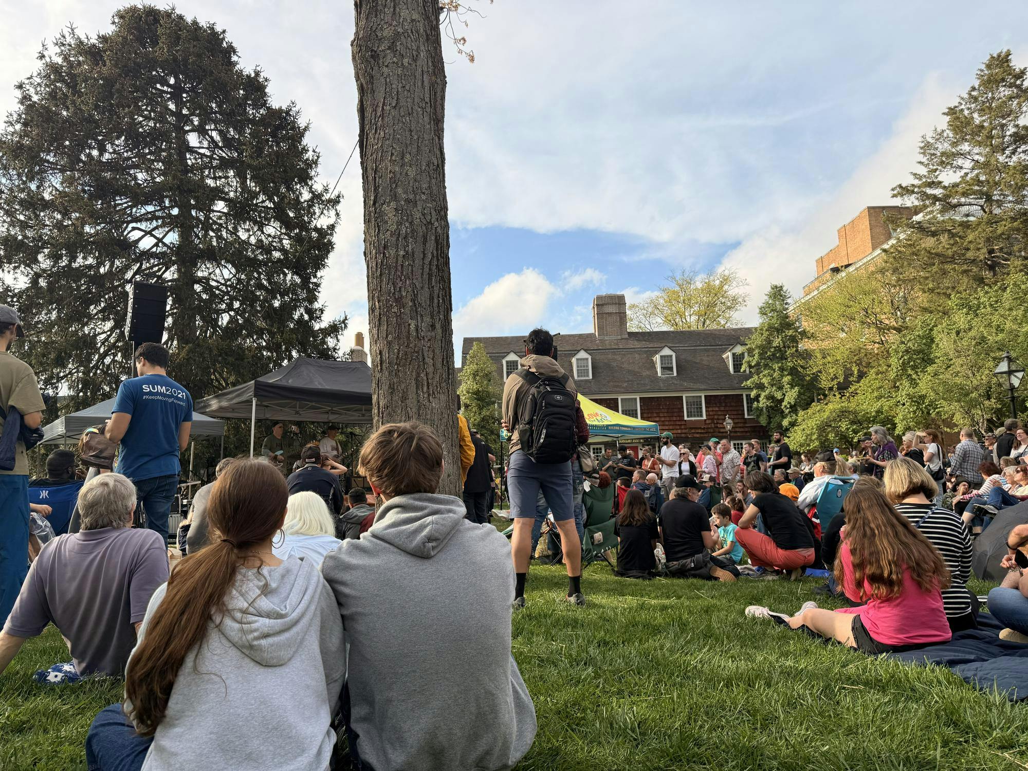 A crowd of students and community members of all ages are gathered on the lawn of a large brick house watching a band perform. 