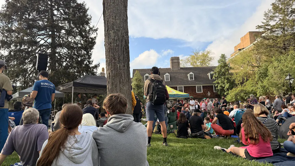 A crowd of students and community members of all ages are gathered on the lawn of a large brick house watching a band perform.