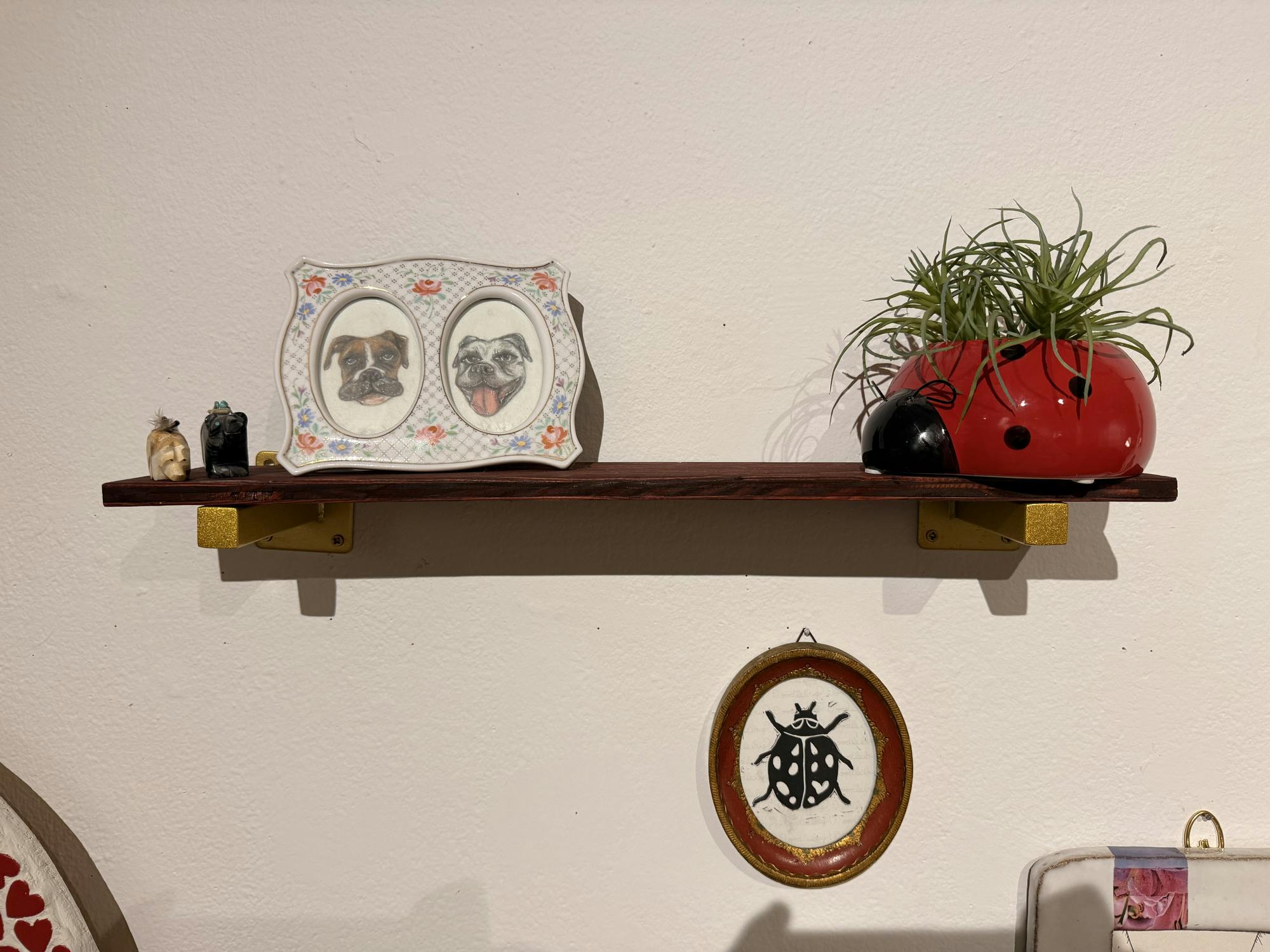 A brown shelf has two small dog-shaped trinkets on the far left, one is beige and one is black. To the right of the trinkets, there is a purple and floral frame that has two pictures of dogs. One of the dogs is brown and one is white with a pink tongue. On the far right of the shelf there is a red ladybug planter with a green plant in it. The brown shelf is hung on a white wall.