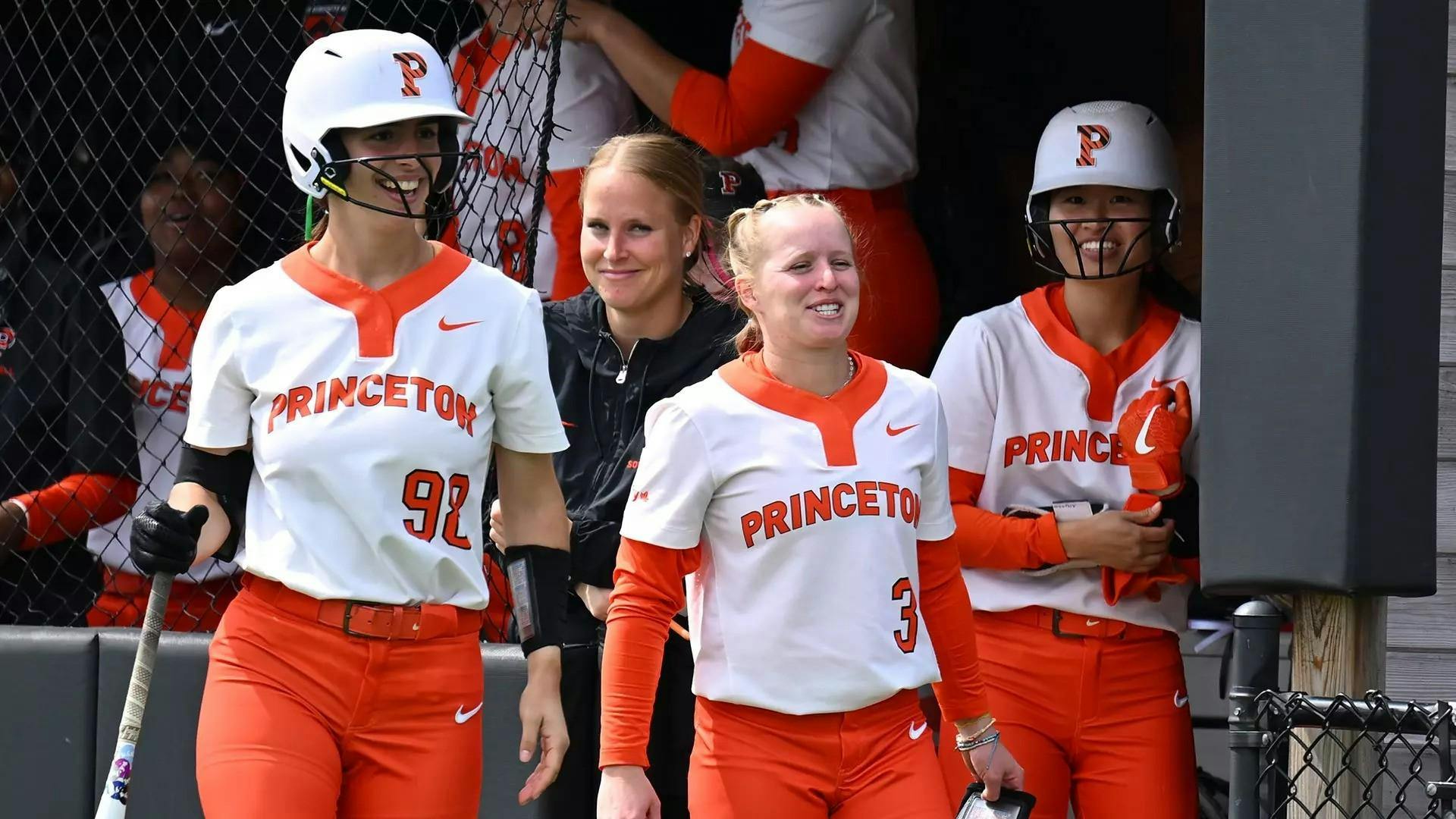 A group of women smiling in the dugout of a baseball field. 