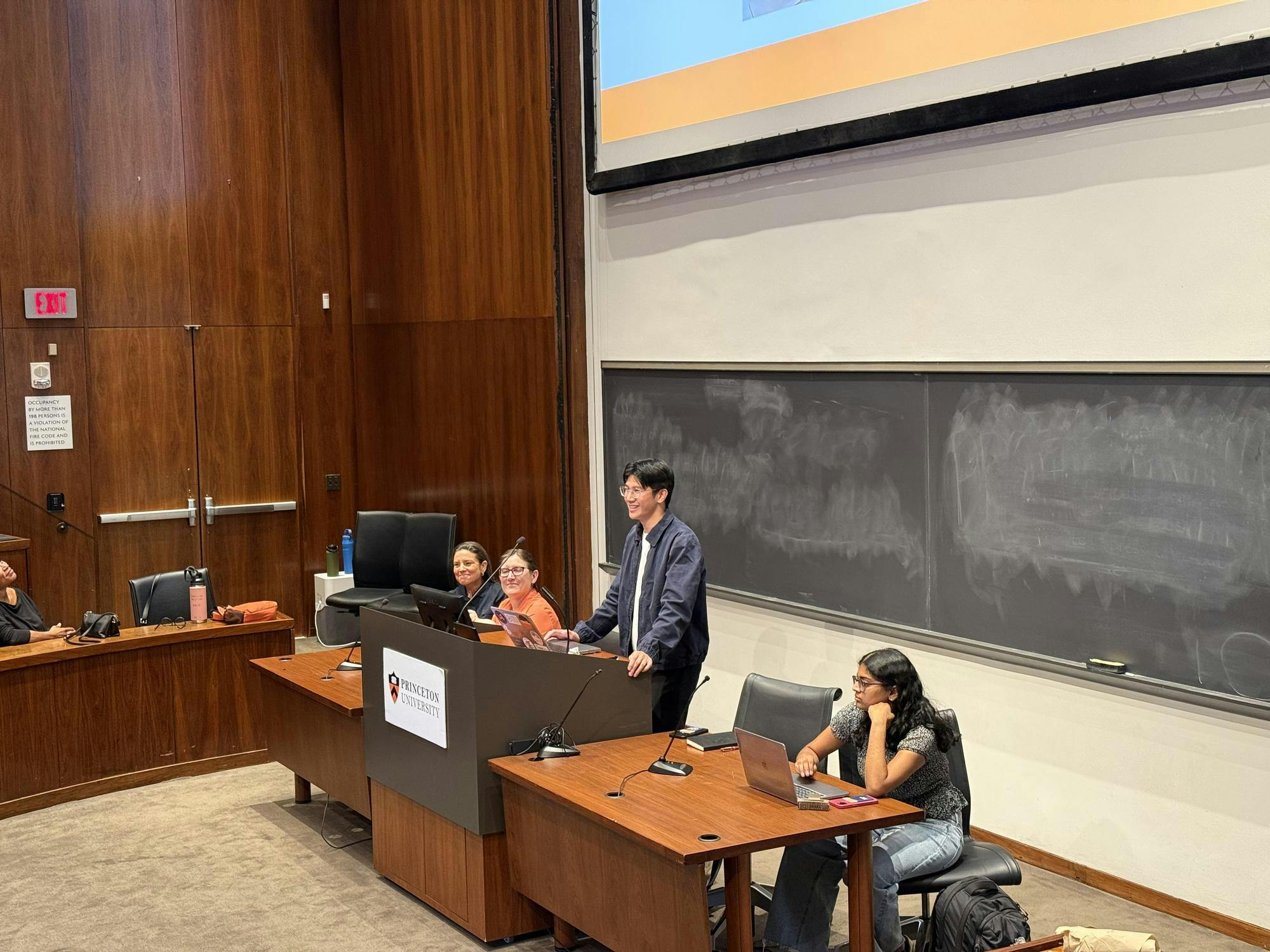 Four people are seated and stood behind a desk in an auditorium. There is a blackboard behind them.