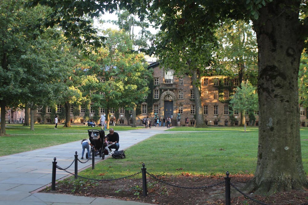 A large brown building, partially obscured by leafy trees, with a person and children gathered around a stroller on the path in front