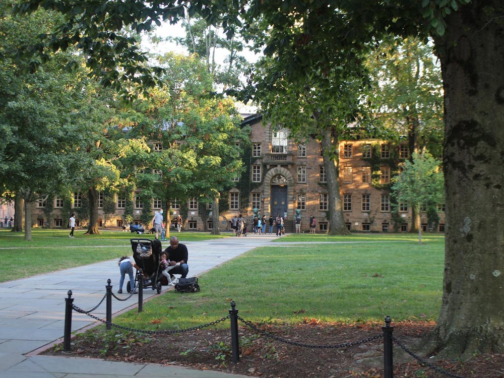A large brown building, partially obscured by leafy trees, with a person and children gathered around a stroller on the path in front