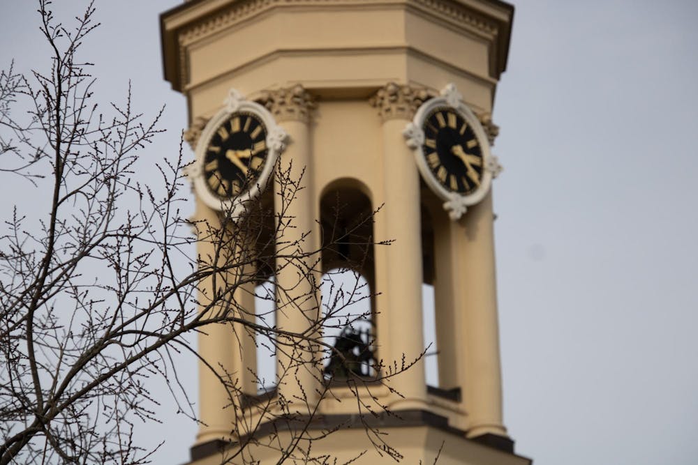 The top of a building with two clocks on the sides in the background of the image, with the branches of a tall tree in the foreground.