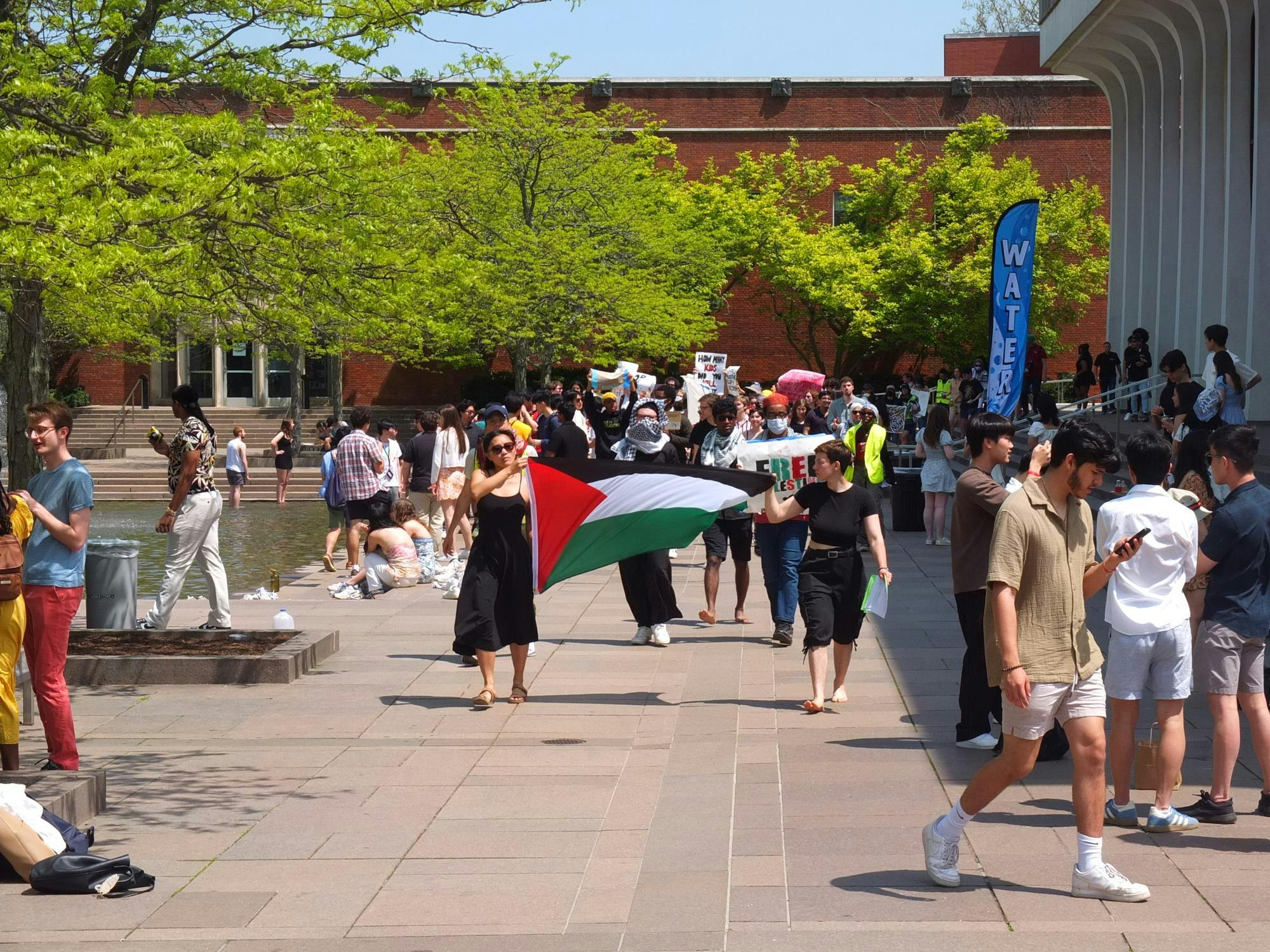 A group of protesters march with a Palestinian flag between them. In the background, students are taking photos. 