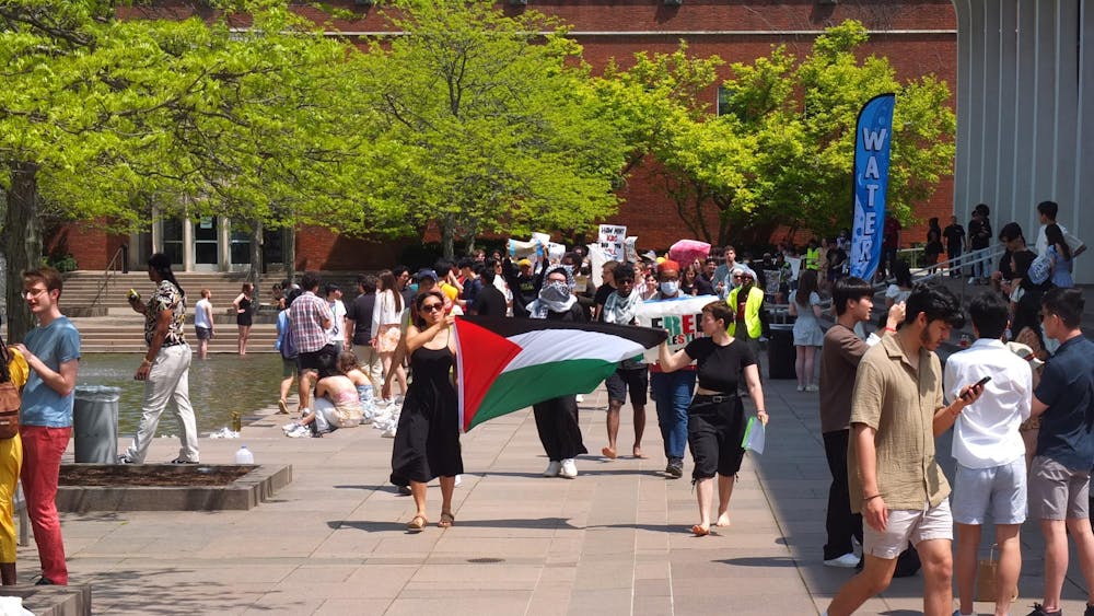 A group of protesters march with a Palestinian flag between them. In the background, students are taking photos. 