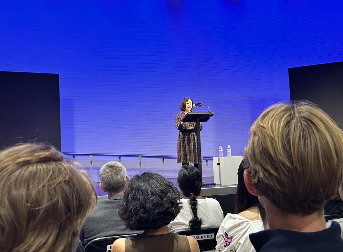 A woman stands at a lectern in a blue-lit room.