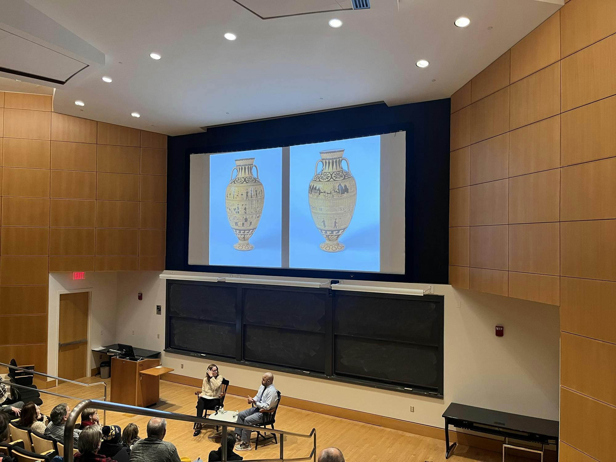 A man in a dress shirt and tie sits in a wooden chair, speaking with a woman in a yellow sweater in a large lecture hall with wooden paneling.  