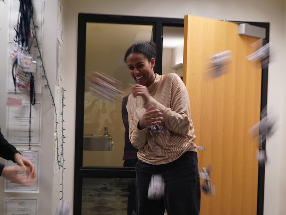 Woman in sweater smilingly braces herself as blurry balls of paper are tossed at her. Door is slightly ajar, name tags hang on hook.