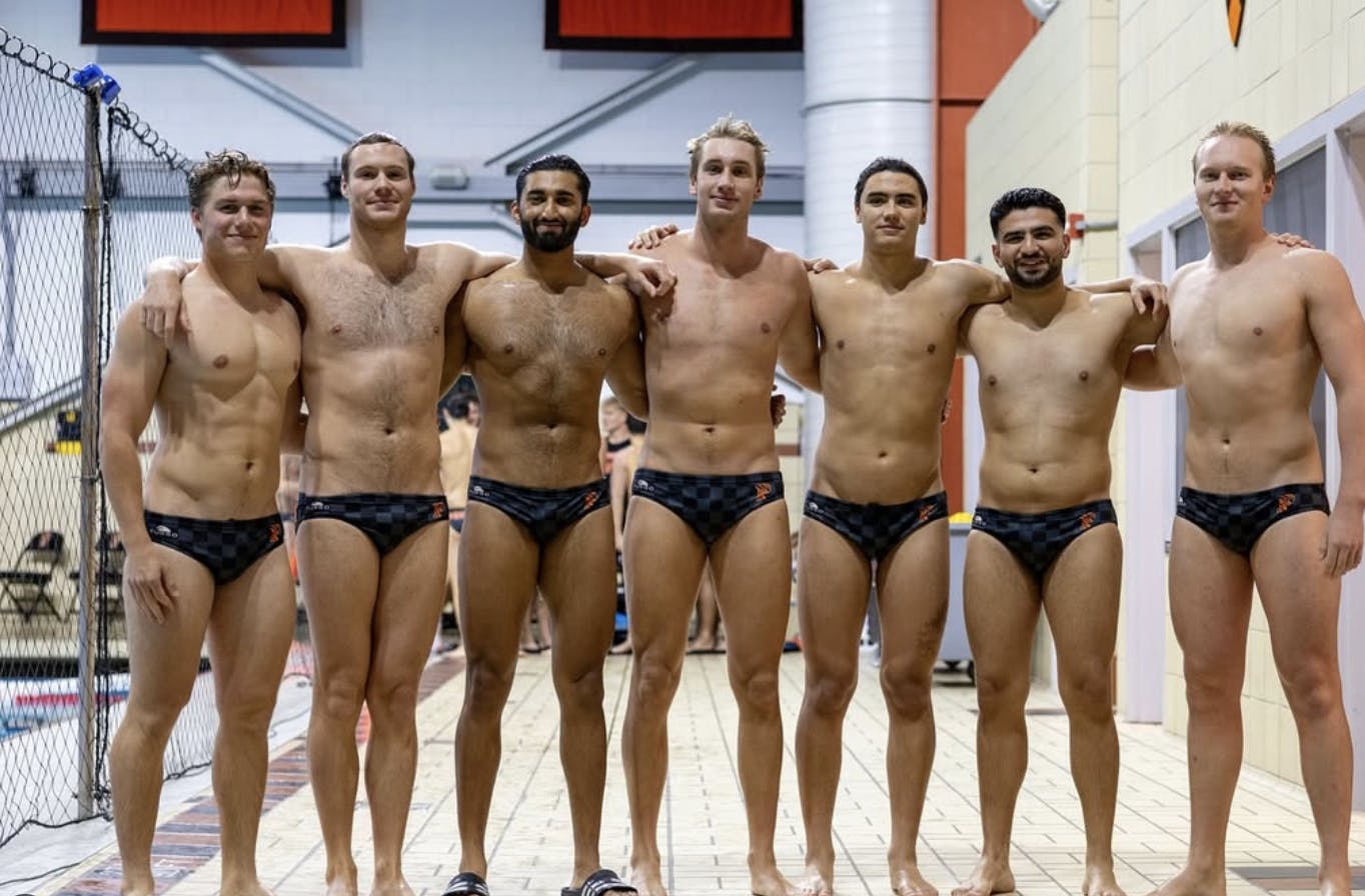 Seven men in black speedos stand in front of a pool