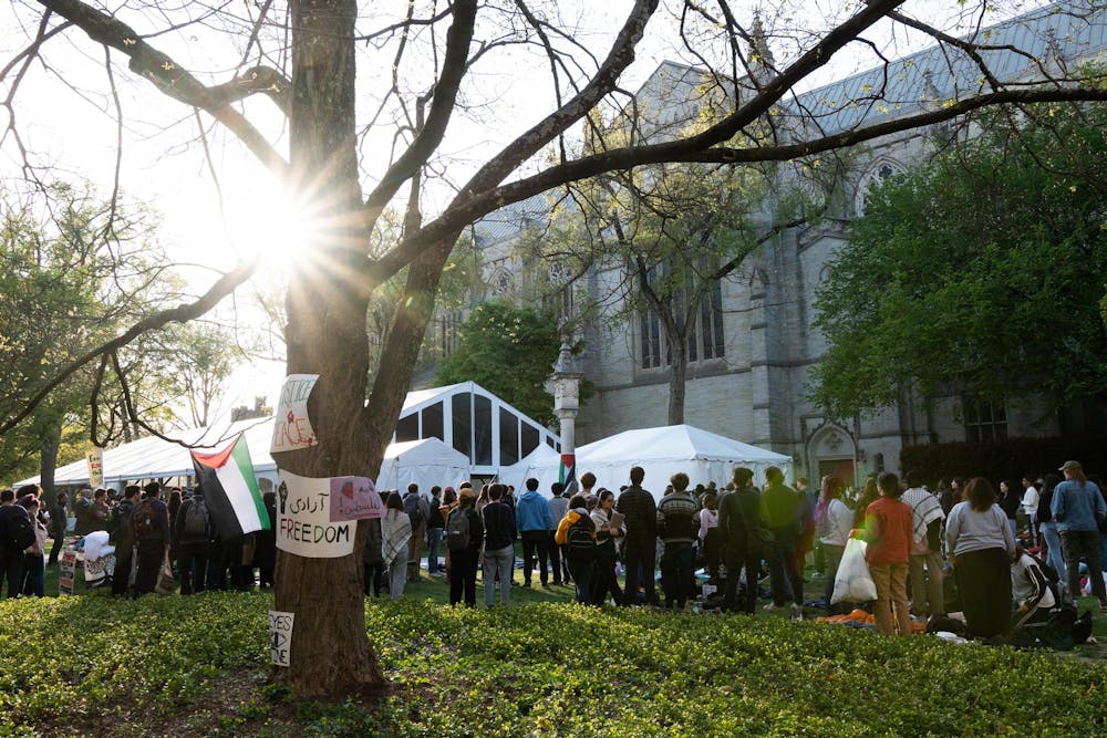 Crowd of people standing, facing away from the camera. In the foreground, a tree with signs reading phrases like “Freedom.”