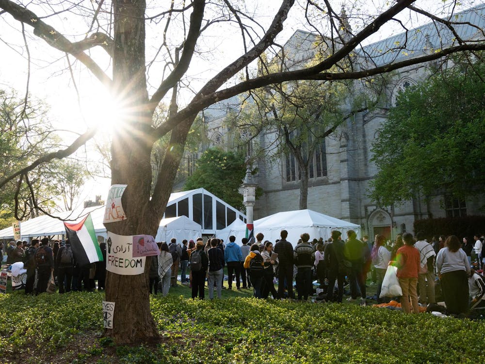 Crowd of people standing, facing away from the camera. In the foreground, a tree with signs reading phrases like "Freedom."