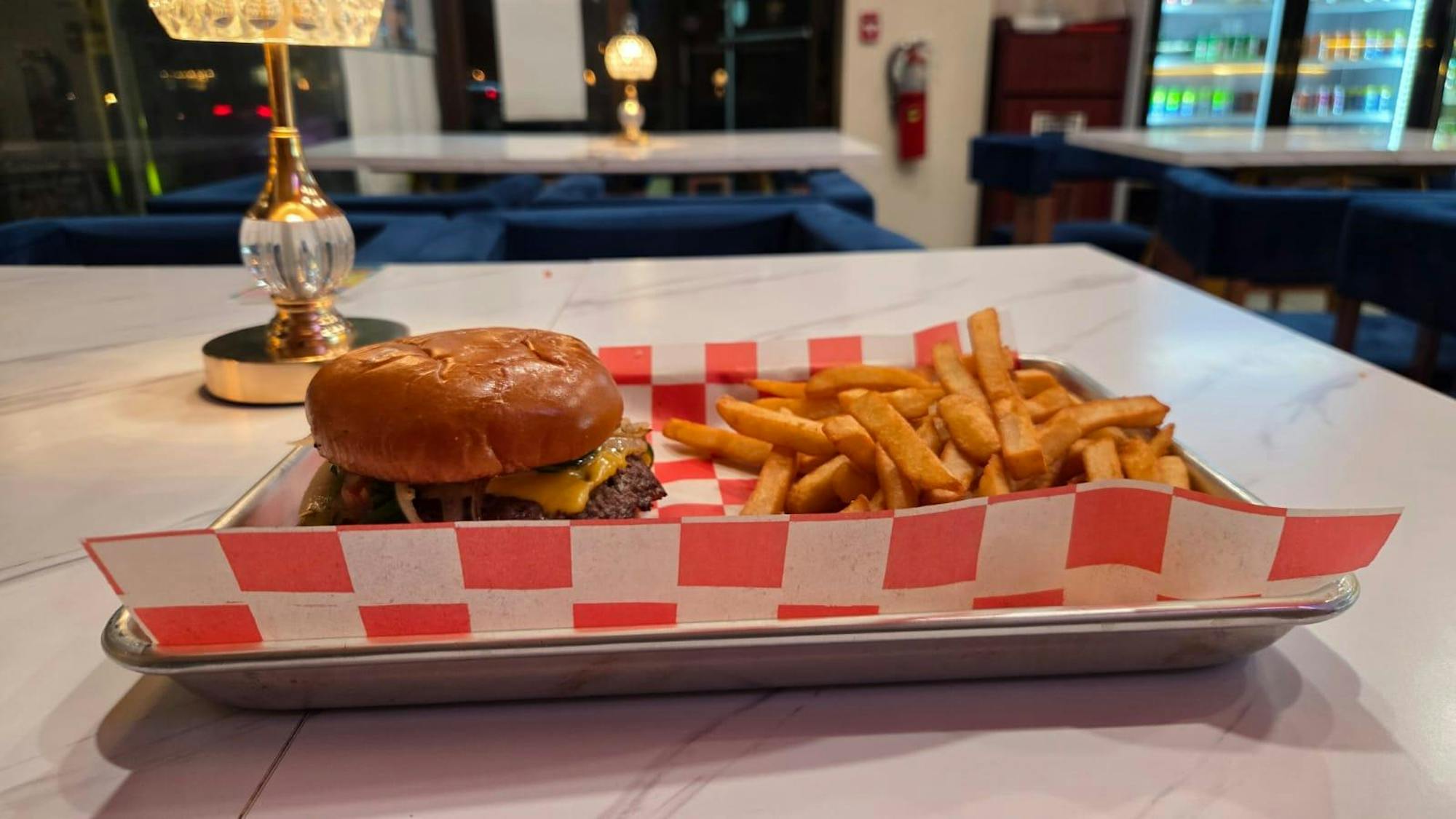 There is a small gray tray with red and white checkered liner on it. There is a brown burger with a black patty and brown fries on the tray. The whole tray sits on a white marbled countertop.