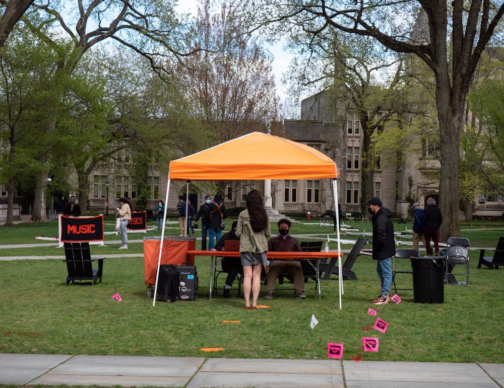 A student faces two people sitting at a table underneath an orange tent. On the lawn, there are concentration banners including Music and Physics.