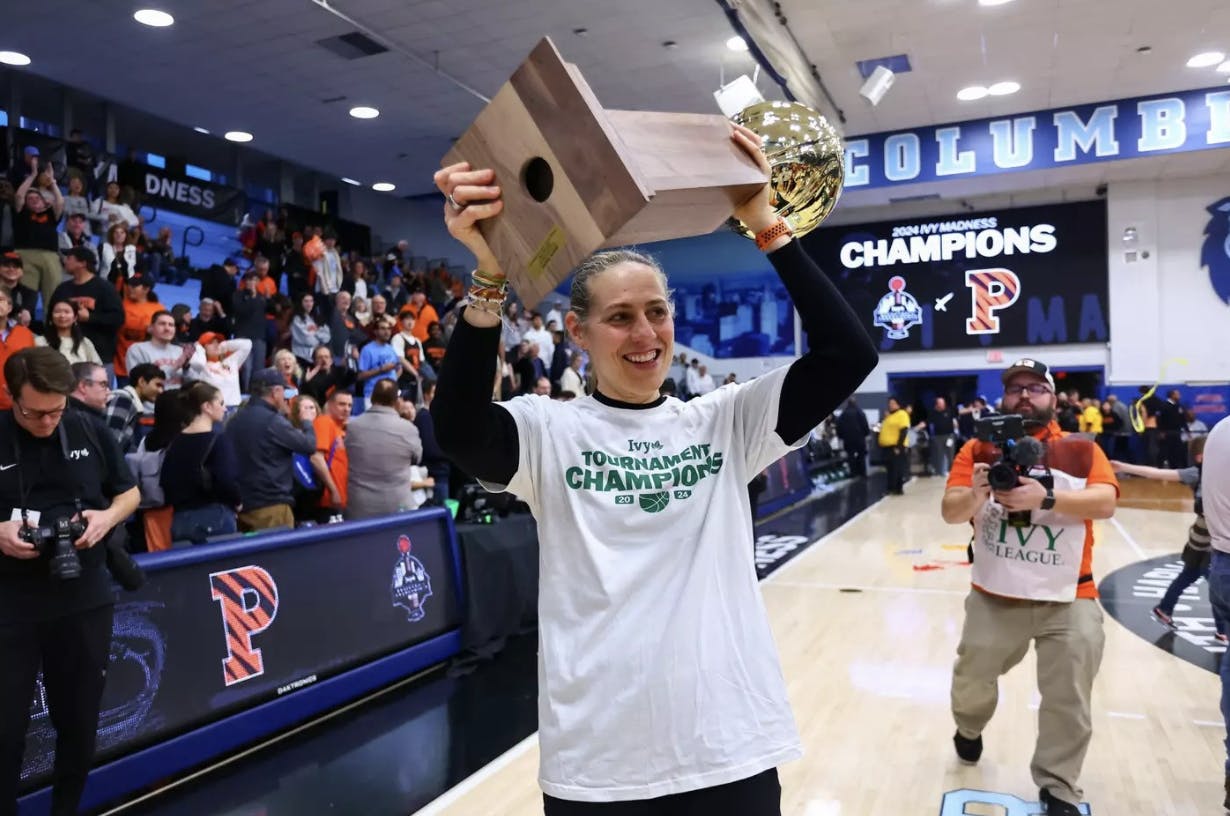 A coach in a white t-shirt lifts a gold trophy and wooden plaque overhead, smiling as a crowd celebrates behind her on a gym court.