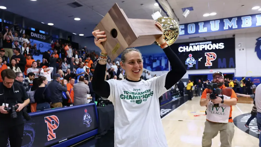 A coach in a white t-shirt lifts a gold trophy and wooden plaque overhead, smiling as a crowd celebrates behind her on a gym court.