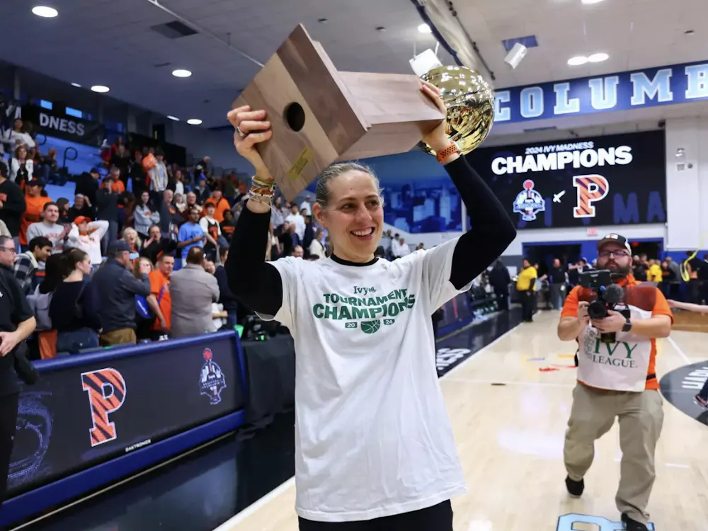 A coach in a white t-shirt lifts a gold trophy and wooden plaque overhead, smiling as a crowd celebrates behind her on a gym court.