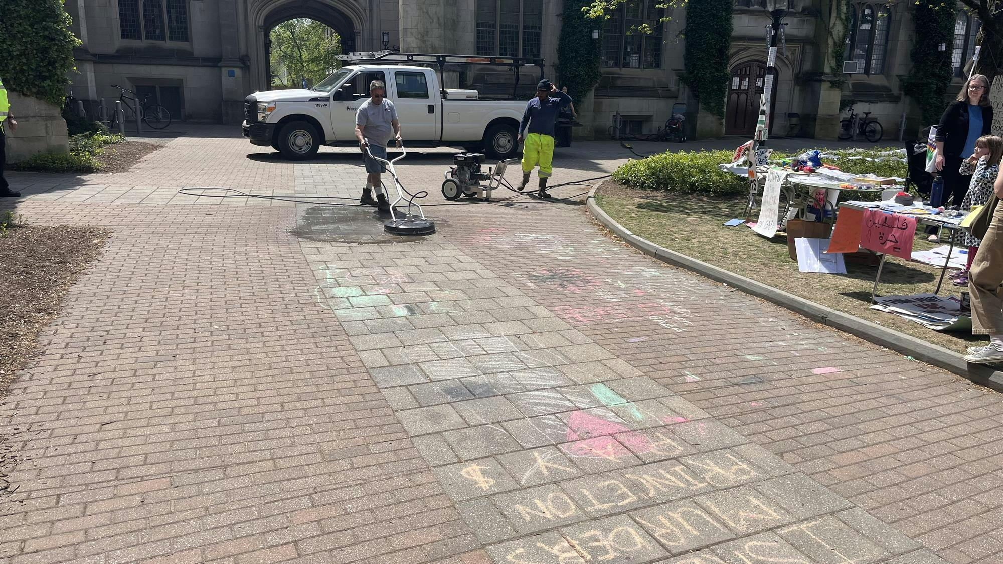 A man in a gray shirt uses a power washing machine to scrub chalk off a sidewalk.