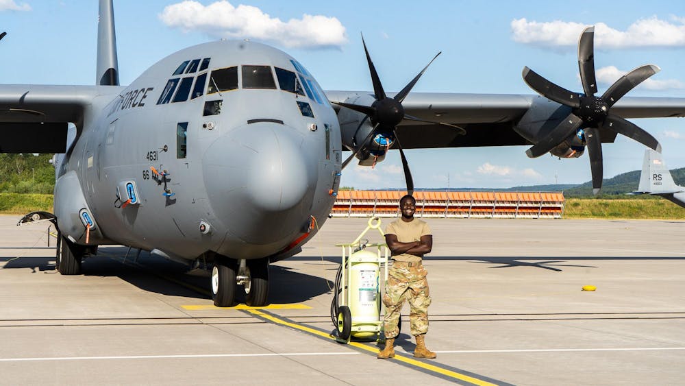 A U.S. Air Force service member stands smiling in front of a C-130 aircraft on a tarmac.
