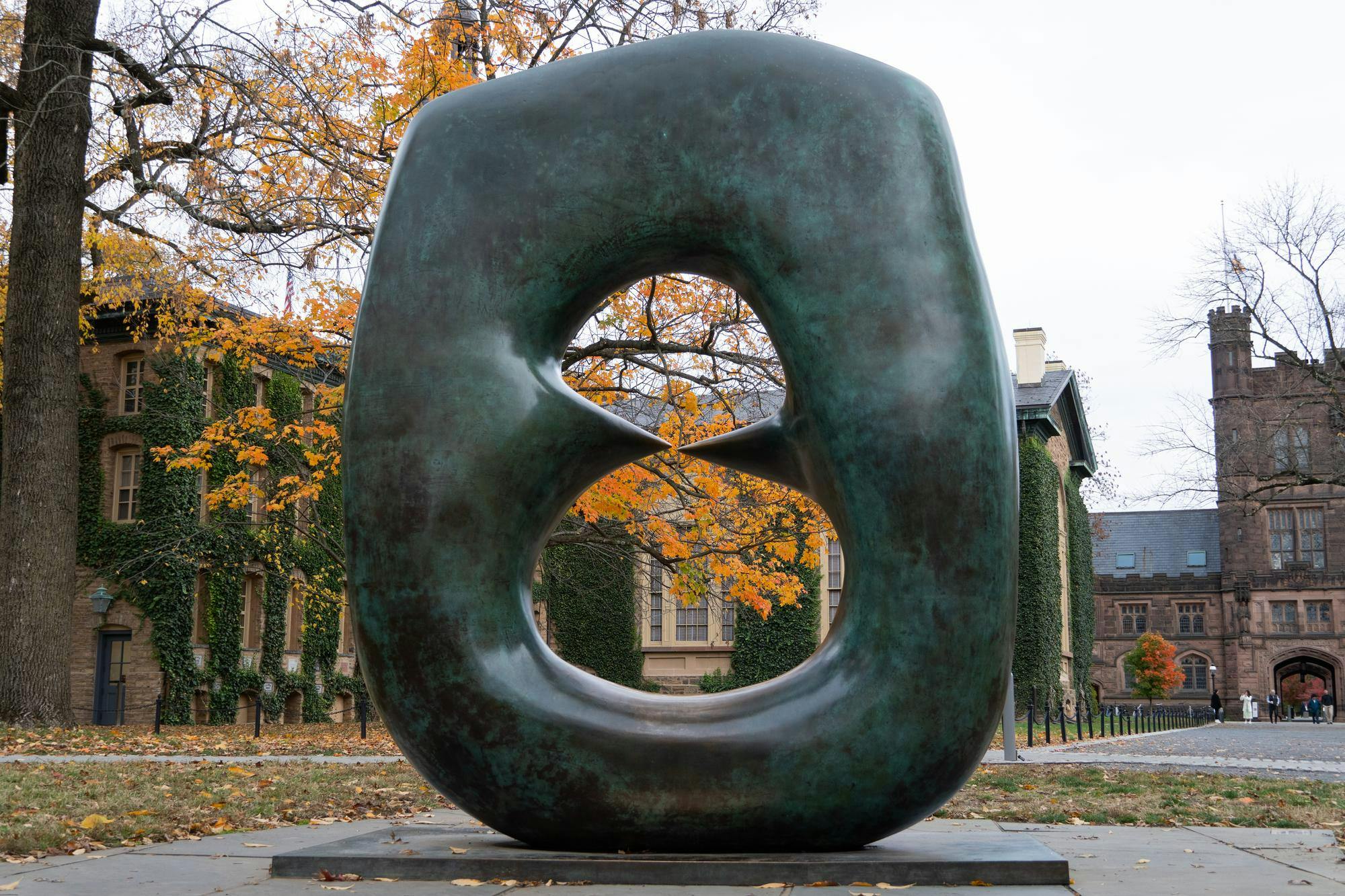 An oval-shaped sculpture, against a backdrop of an ivy-covered building and a tree with yellow leaves
