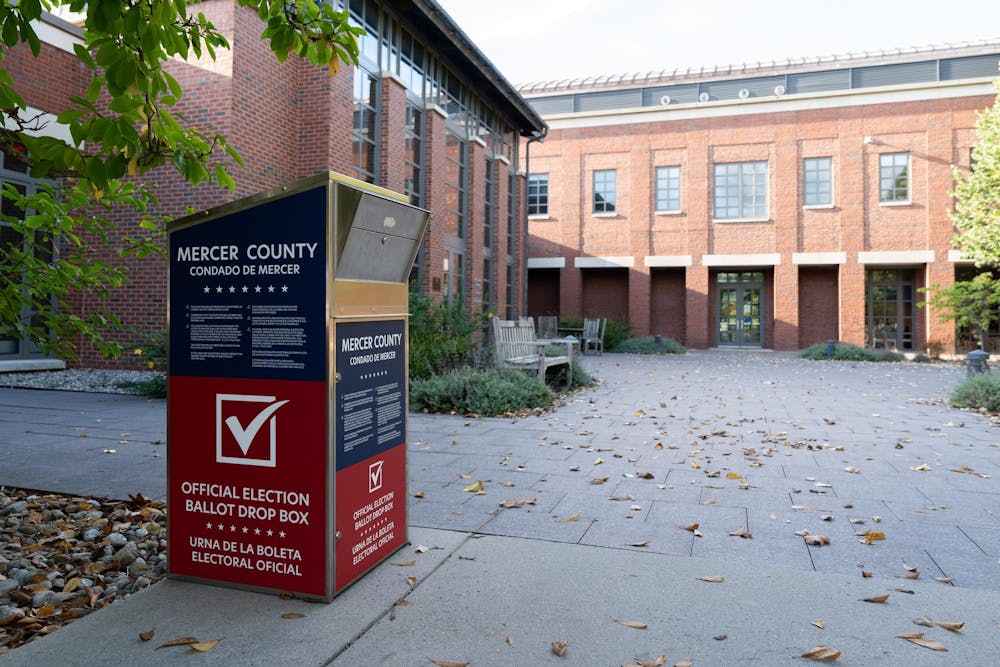 A blue and red box with text reading "official election ballot drop box" in front of a brick building on a fall day.